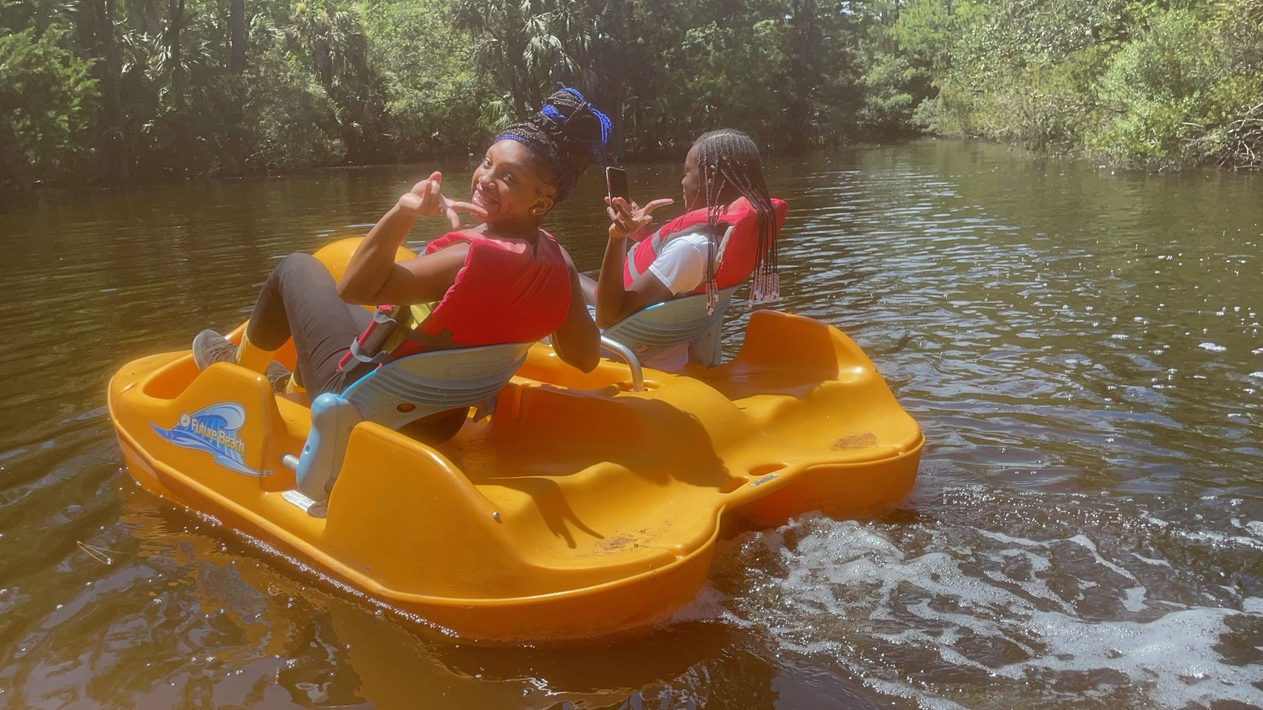 Two girls pose for a picture while pedal boating on a lake.