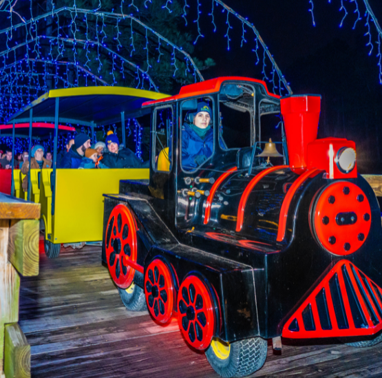 A train drives on a wooden pathway through holiday lights.
