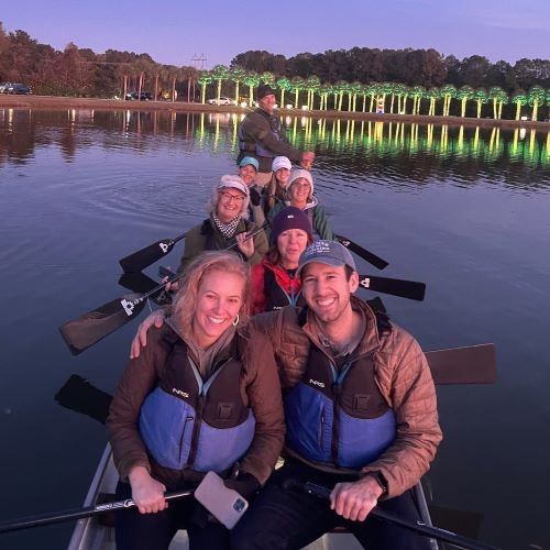 People smiling from a dragon boat while paddling on the lake with bright holiday lights behind them