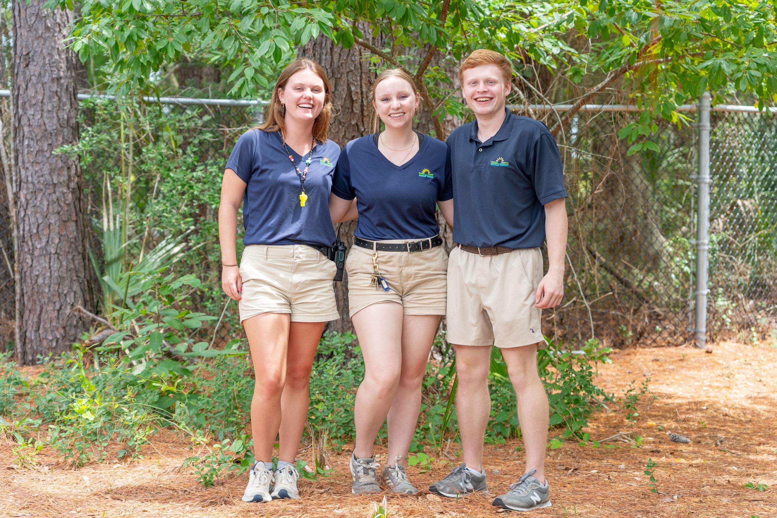 Three interns, two women and a man, standing on pinestraw. 